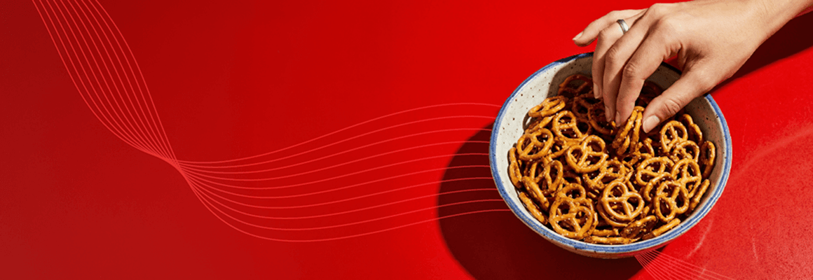 A hand reaching into a ceramic bowl filled with mini pretzels on a bold red background with decorative white lines.
